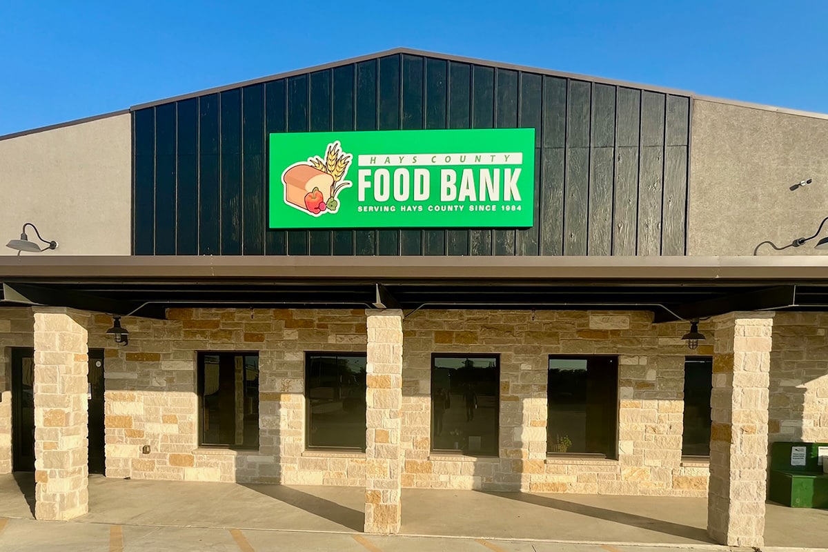 Exterior view of the Hays County Food Bank building featuring a green sign with the text "HAYS COUNTY FOOD BANK" and an illustration of bread and plants, situated under a clear blue sky.