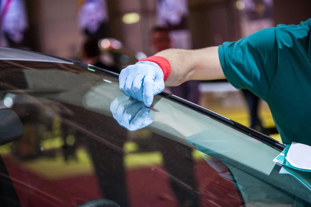 A person wearing a blue glove and a red wristband is using a small tool to examine or repair the windshield of a car.