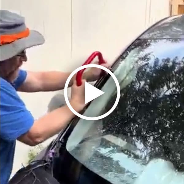 A man wearing a hat and blue shirt is using a red tool to install or remove a windshield on a vehicle, with a blurred background of a wall and trees. A protective black cover is placed over part of the vehicle.