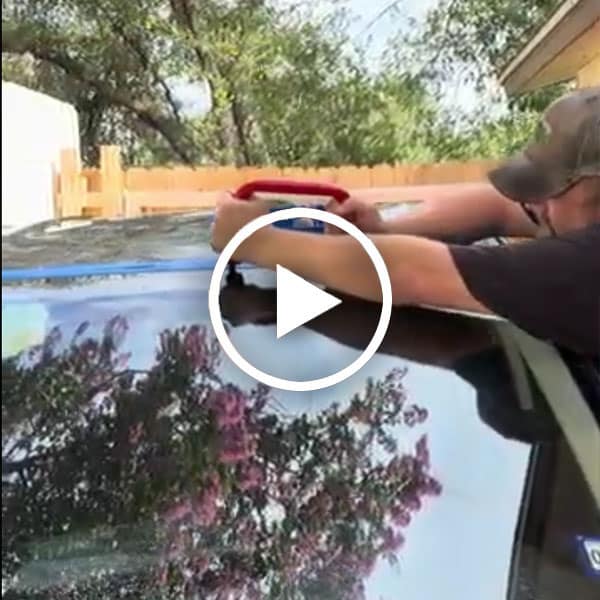 A person applies a tool to the top of a car, with trees and a wooden fence in the background, reflecting on the car's surface.