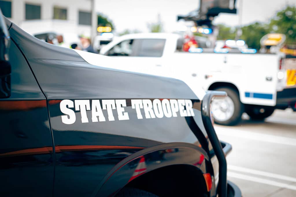 Close-up of a black vehicle featuring the words "STATE TROOPER" in bold white letters on its side, with emergency vehicles and people blurred in the background.