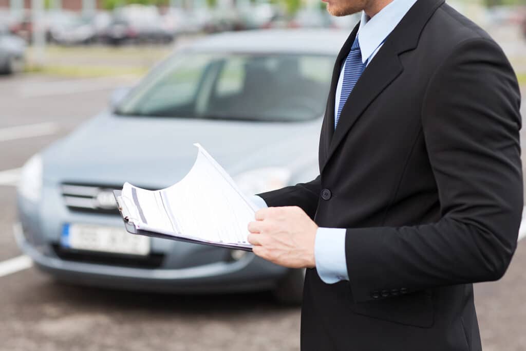 A man in a black suit holds a clipboard and reviews paperwork in front of a silver car parked outdoors.