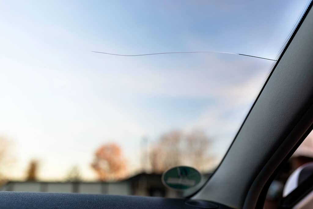 A long, thin horizontal crack extending across a vehicle's windshield, viewed from inside the cabin looking out toward a blue sky and blurred outdoor trees.