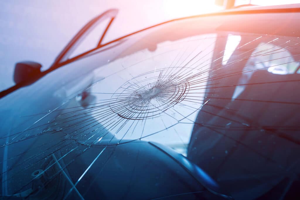 Close-up of a severely cracked car windshield showing a large bullseye impact point with spiderweb fractures spreading across the glass.