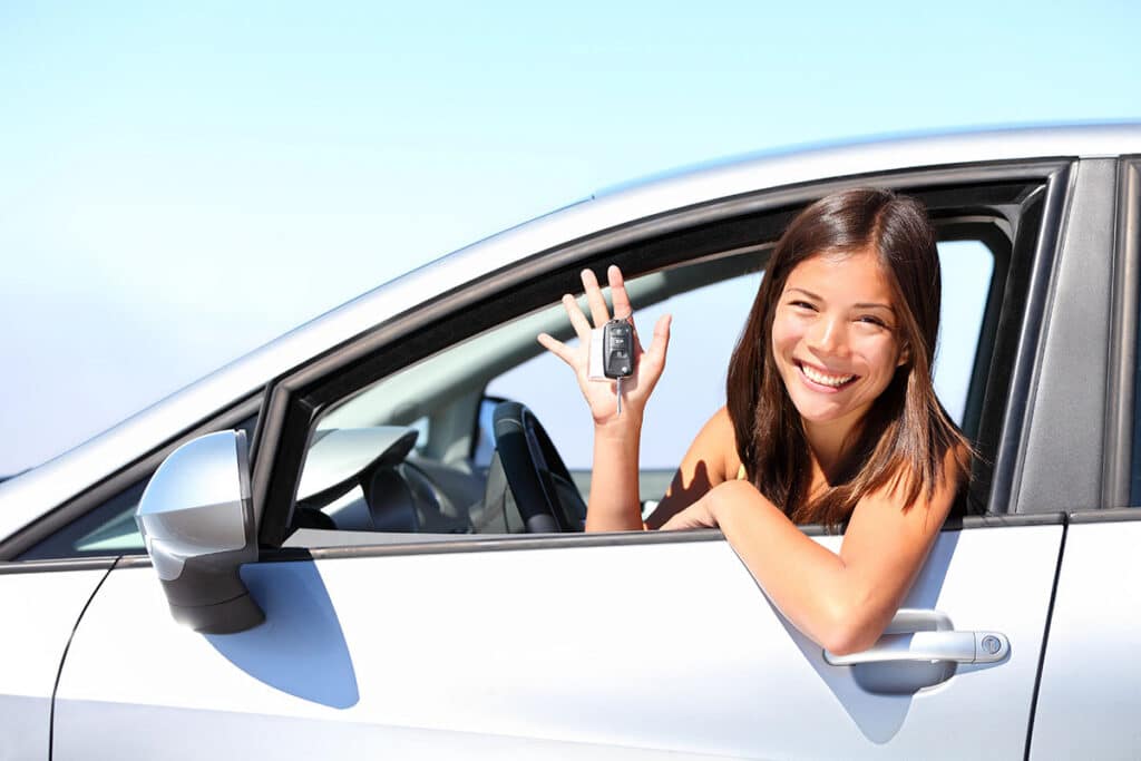 A smiling young woman leaning out of a silver car's driver-side window, holding up a car key fob.