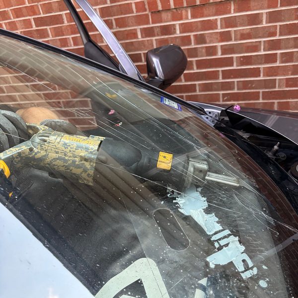 A close-up view of a car's interior, showing a damaged windshield with scratches and a section covered in white adhesive. A hand holding a tool is visible, and part of the car door is open against a brick wall in the background.