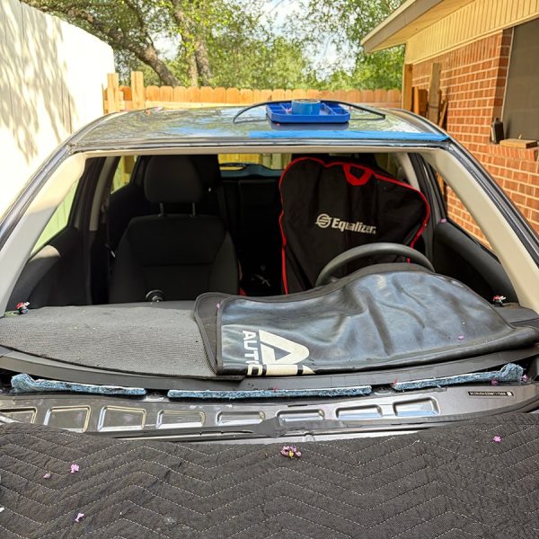 Interior view of a car with a missing rear windshield, displaying debris and a protective cover on the back seat, set against a background of trees and a wooden fence.