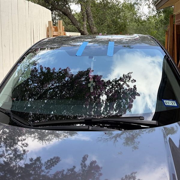 A car windshield with blue painter's tape placed in two horizontal stripes at the top. Reflected in the glass are trees and clouds. A wooden structure is visible in the background, along with a light-colored fence.
